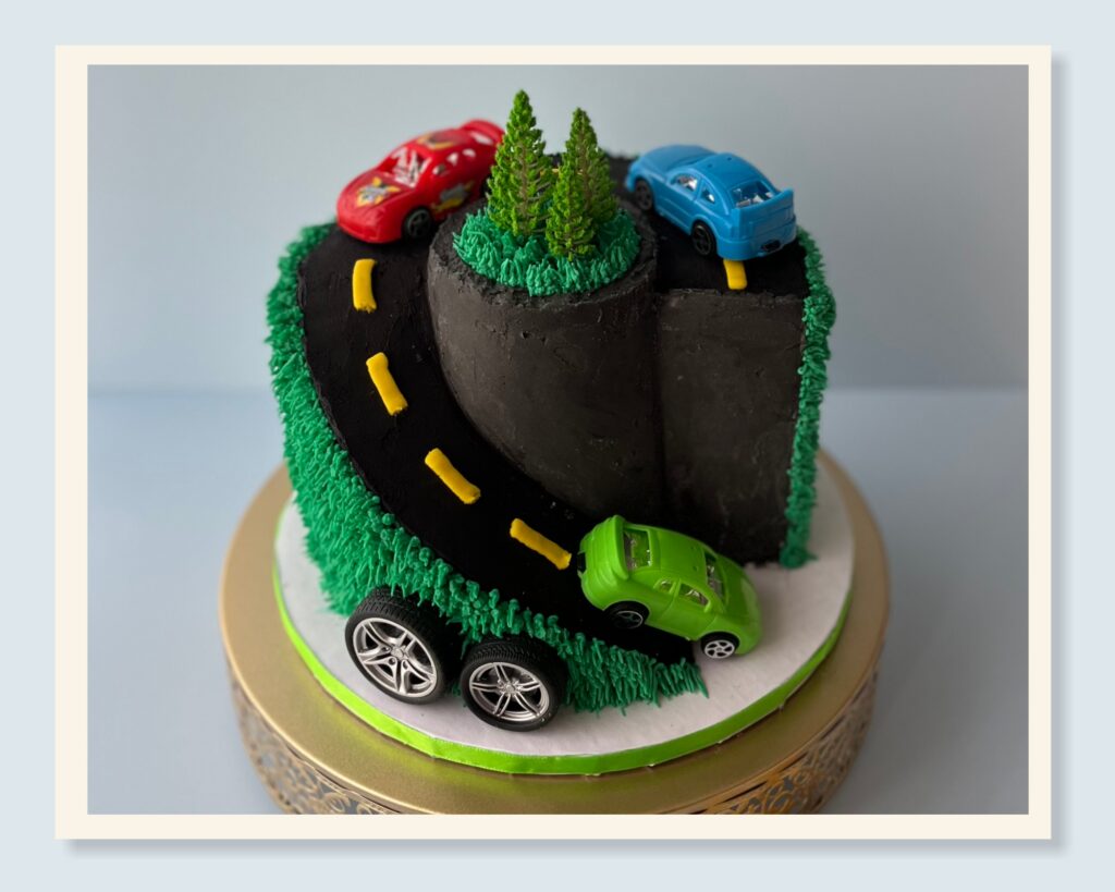 Custom carved car road cake with winding roadway design, toy cars, and textured green buttercream grass