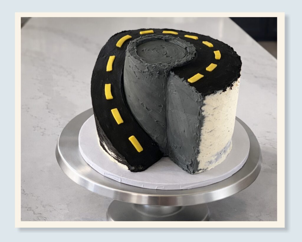 Partially finished custom cake showing the cake design process, with textured buttercream grass and road details being added.
